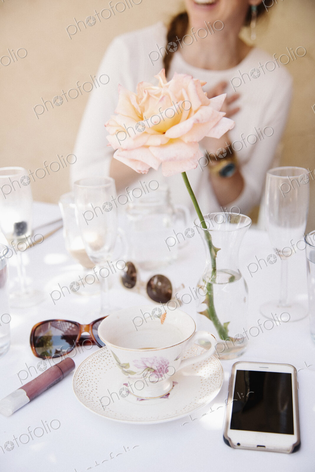 Close up of a rose in a vase on a table with cups and glasses, a cell phone and…