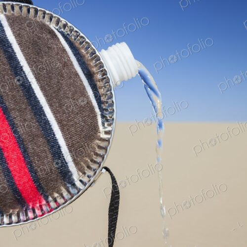 Close-up of a striped fabric-covered canteen tilted to pour out a stream of water,…