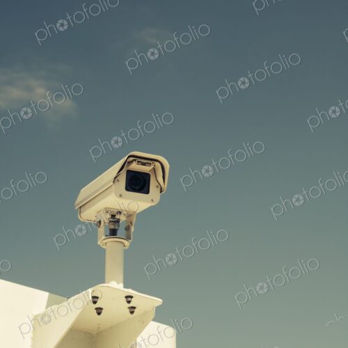Close-up of a surveillance camera mounted outdoors against a clear blue sky,… Close-up of a surveillance camera mounted outdoors against a clear blue sky,…