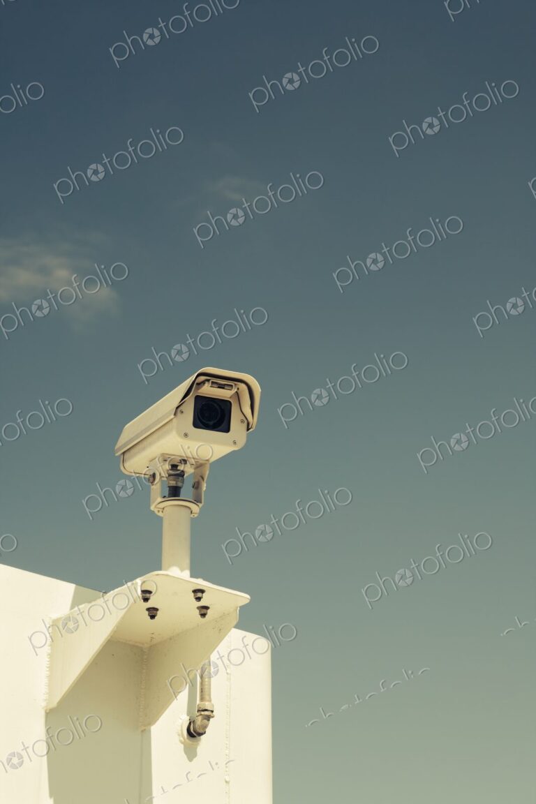 Close-up of a surveillance camera mounted outdoors against a clear blue sky,…