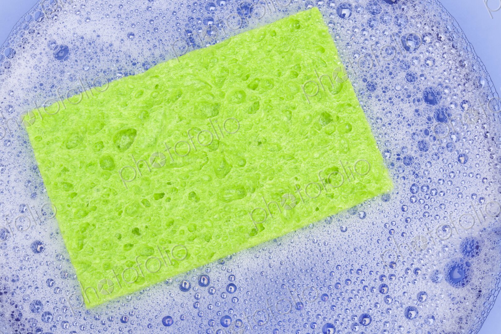 Close-up of a vibrant green sponge resting in a bowl of foamy soapy water. The image…