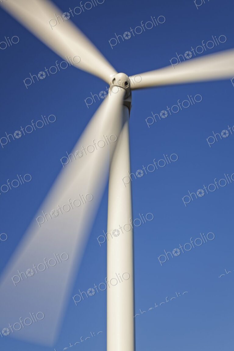 Close-up of a wind turbine with spinning blades against a clear blue sky, symbolizing…
