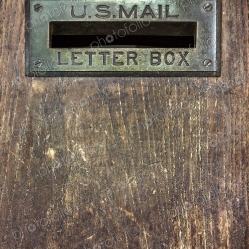 Close up of an antique U.S. Mail letter slot set in a worn wooden door, showing brass…