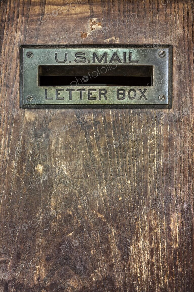 Close up of an antique U.S. Mail letter slot set in a worn wooden door, showing brass…