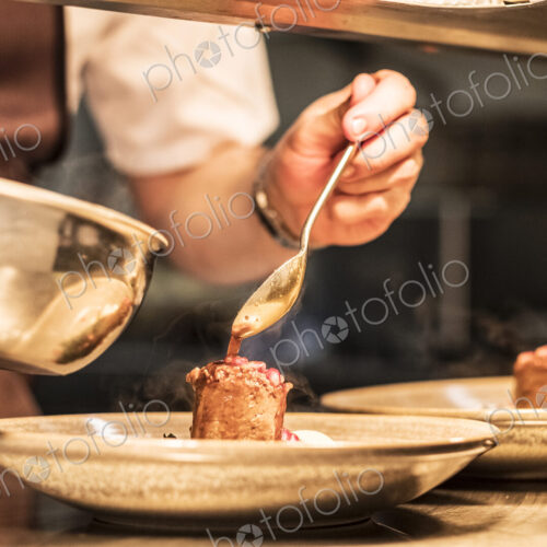 Close up of chef in commercial kitchen holding spoon, drizzling sauce on plate.