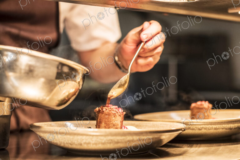Close up of chef in commercial kitchen holding spoon, drizzling sauce on plate.