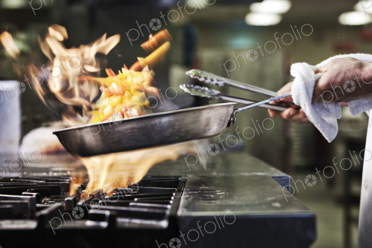 Close-up of chef's hands holding a sauté pan to cook food, flambeing contents. Flames…