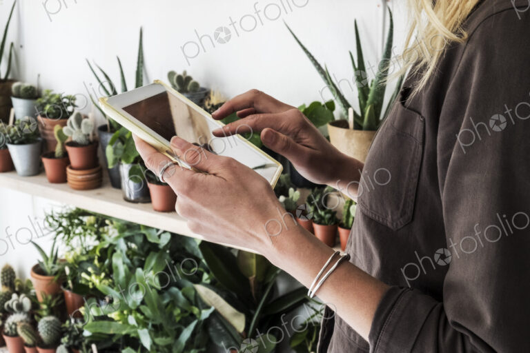 Close up of female owner of plant shop holding digital tablet, a selection of plants…