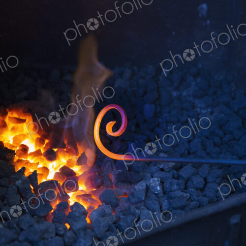 Close up of glowing metal rod with a scrolled end, held over hot coals in a…