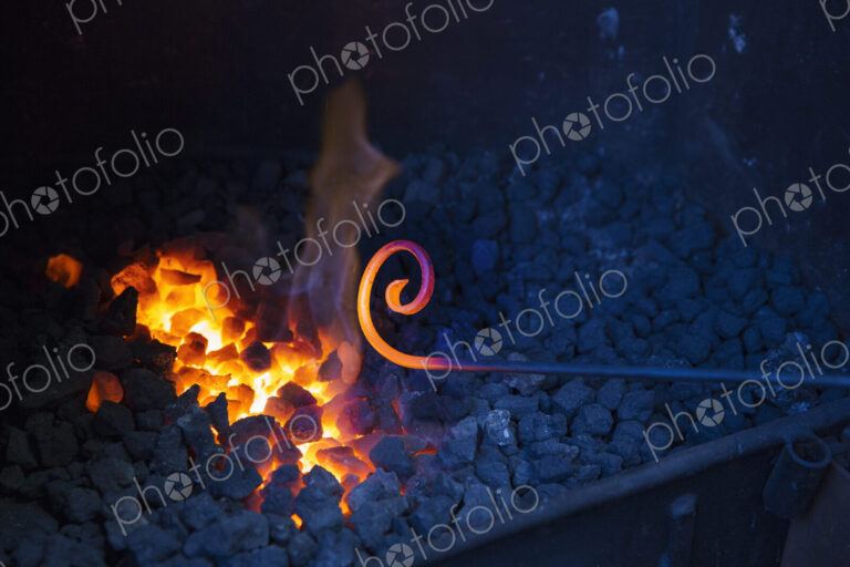 Close up of glowing metal rod with a scrolled end, held over hot coals in a…