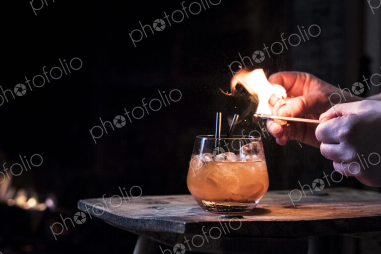 Close up of human hand striking match over glass with alcoholic drink.