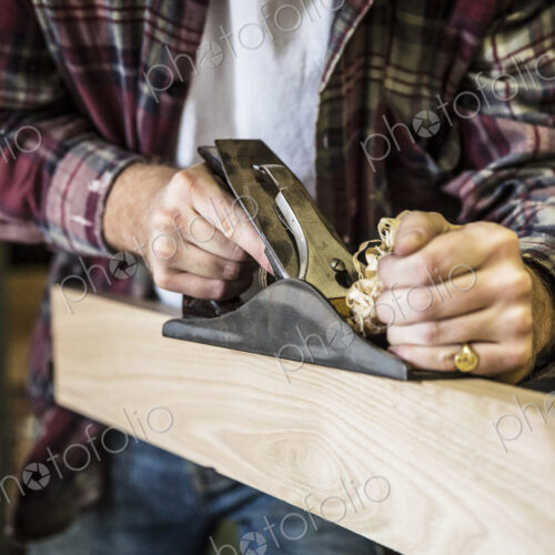 Close up of man working standing in a woodworking workshop, using plane on plank of wood.
