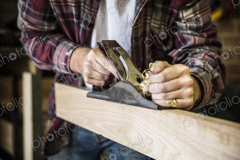 Close up of man working standing in a woodworking workshop, using plane on plank of wood.