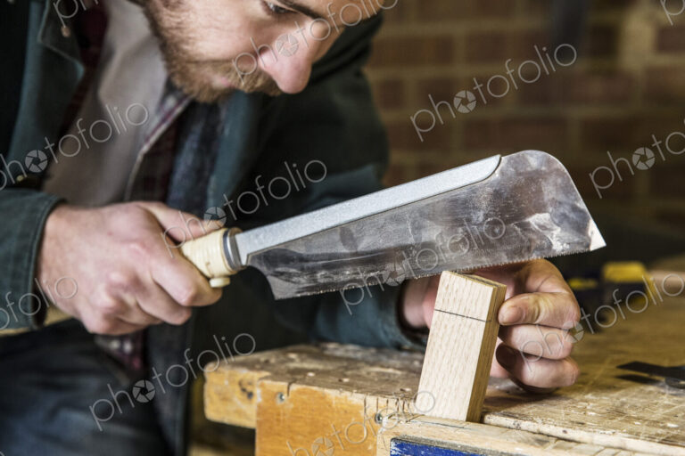 Close up of man working standing in a woodworking workshop, using saw on piece of wood.