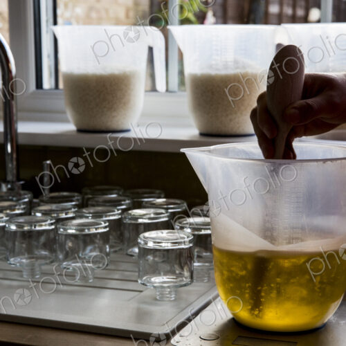 Close up of person holding jug, stirring liquid wax for candle making with wooden spoon.