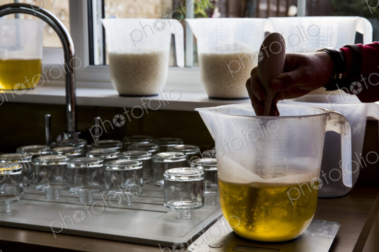 Close up of person holding jug, stirring liquid wax for candle making with wooden spoon.