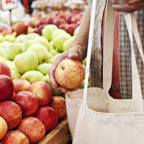 Close up of person holding shopping bag and red apple at a fruit and vegetable market.
