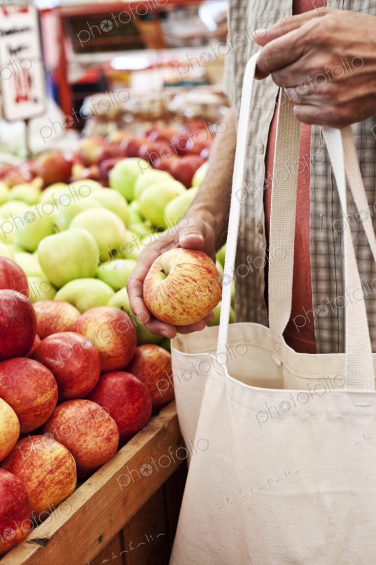 Close up of person holding shopping bag and red apple at a fruit and vegetable market.