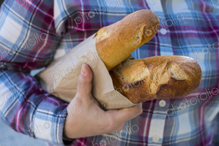 Close up of person holding two freshly baked French baguettes in brown paper bag.