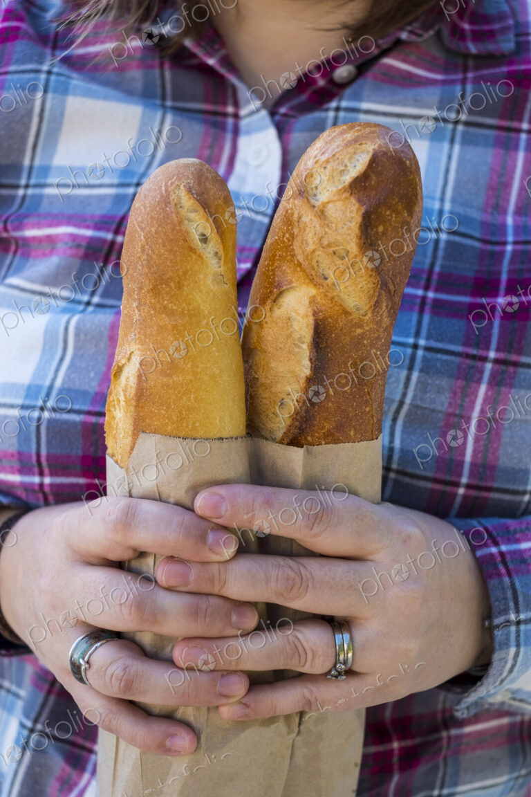 Close up of person holding two freshly baked French baguettes in brown paper bag.