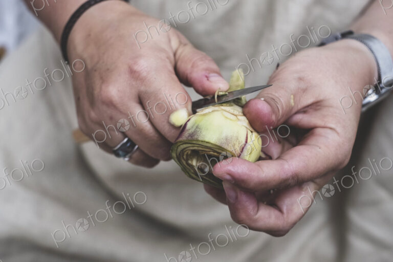 Close up of person peeling fresh artichoke with kitchen knife.