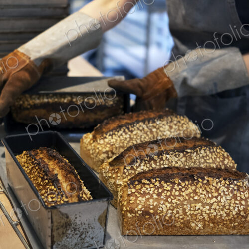 Close up of person wearing oven gloves placing freshly baked seeded loaves of bread on…