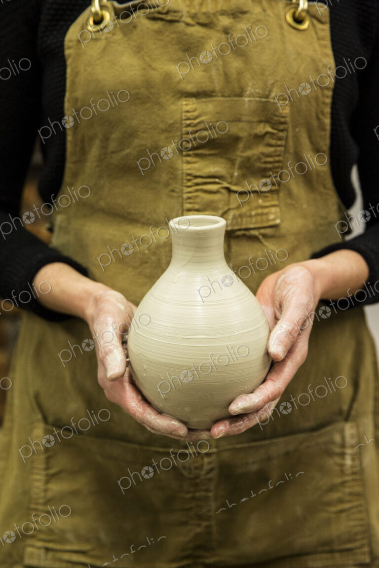 Close up of potter wearing apron holding unfired spherical clay vase.