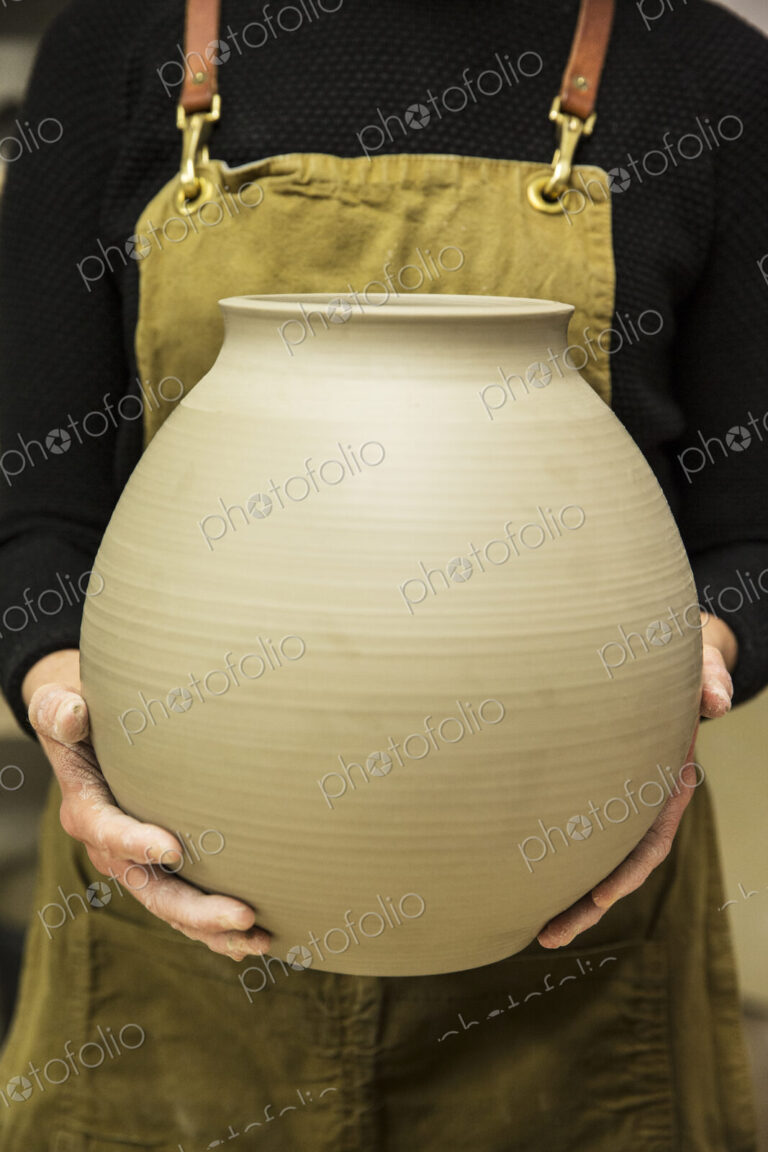 Close up of potter wearing apron holding unfired spherical clay vase.