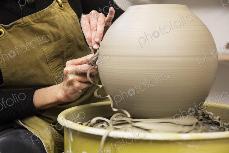 Close up of potter wearing apron working on spherical clay vase on pottery wheel.