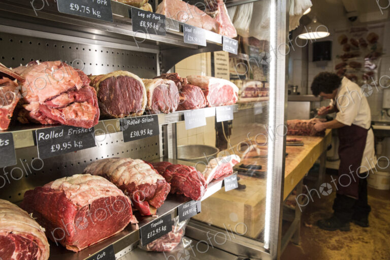 Close up of various cuts of beef on shelves in a butcher shop, man wearing apron…