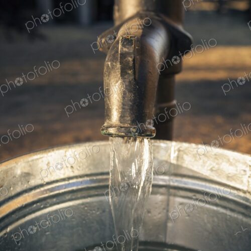 Close-up of water flowing from an old metal pump into a galvanized steel bucket. Rural…