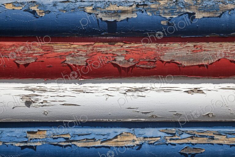 Close-up of weathered red, white, and blue paint peeling off wooden boards, evoking…