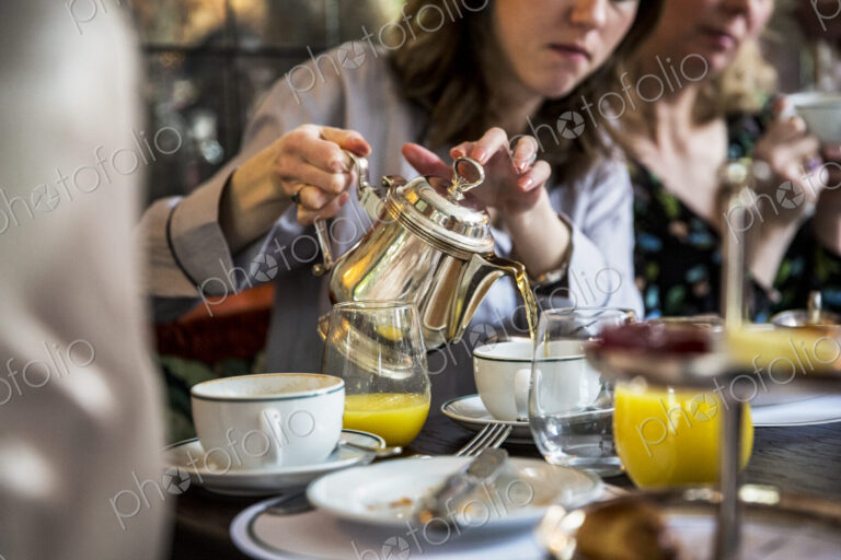 Close up of woman sitting at a table, pouring tea from silver tea pot.