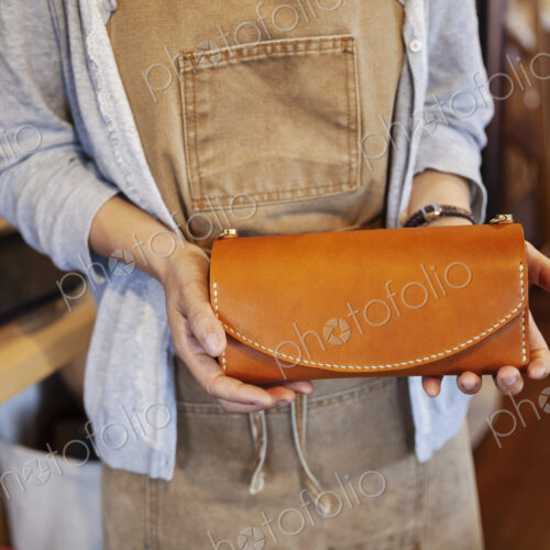 Close up of woman standing in a leather shop, holding tan leather clutch bag.