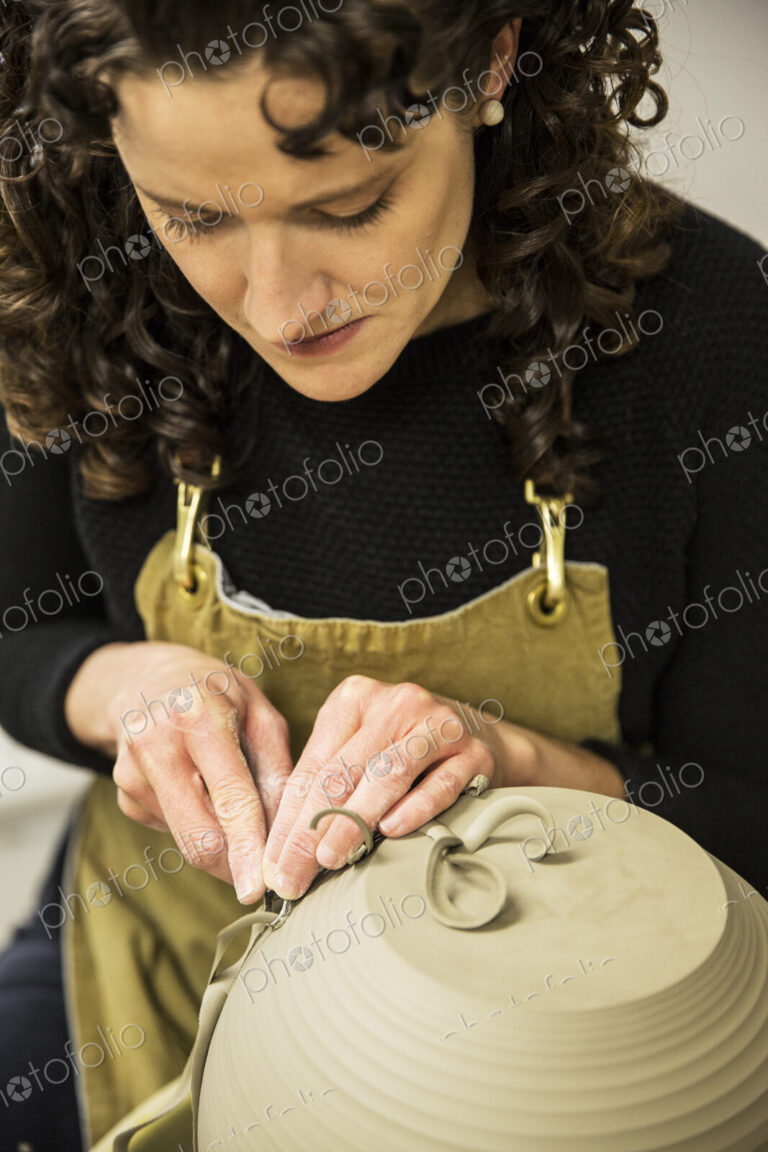 Close up of woman with curly brown hair wearing apron working on spherical clay vase…