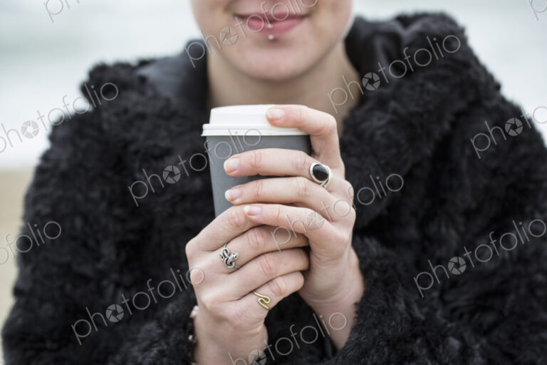 Close up of young woman wearing black furry jacket holding paper cup with hot drink.