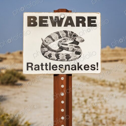 Close view of a Beware Rattlesnakes sign against blue sky and dry prairie in Badlands… Close view of a Beware Rattlesnakes sign against blue sky and dry prairie in Badlands…