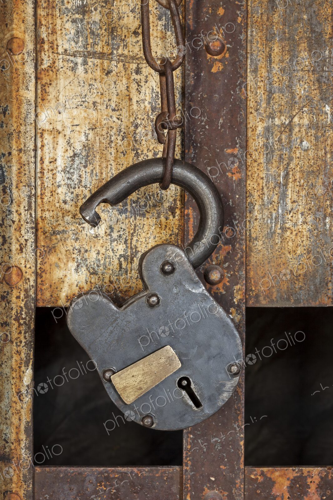 Close view of a vintage padlock and chain hanging on a rusty jail door, with iron bars…