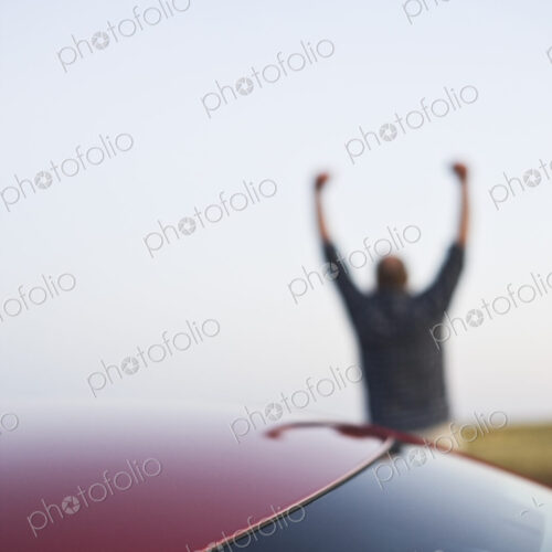 Closeup of a car window and a single male celebrating with his arms up in the background.