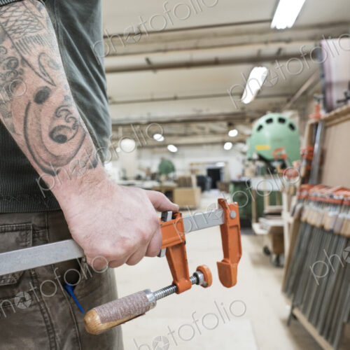 Closeup of an arm with tatoos and a hand holding a bar clamp in a large woodworking… Closeup of an arm with tatoos and a hand holding a bar clamp in a large woodworking…