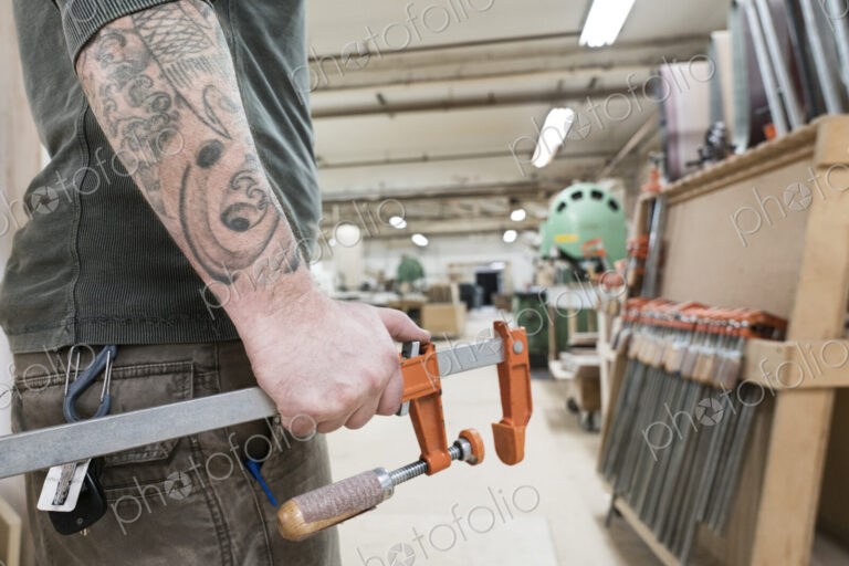 Closeup of an arm with tatoos and a hand holding a bar clamp in a large woodworking…