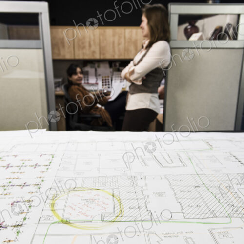 Closeup of plans on a table with two people working in the background.