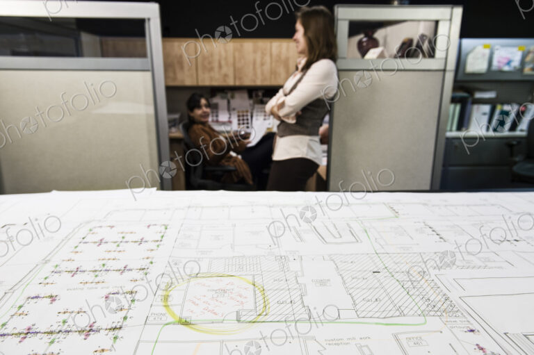 Closeup of plans on a table with two people working in the background.