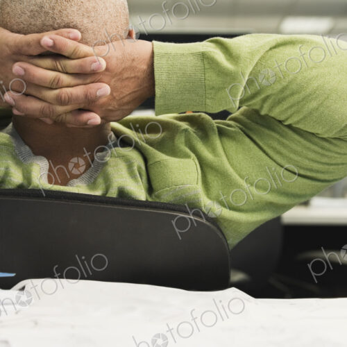 Closeup view from behind of a Black man in a cubicle office with hands behind his head. Closeup view from behind of a Black man in a cubicle office with hands behind his head.