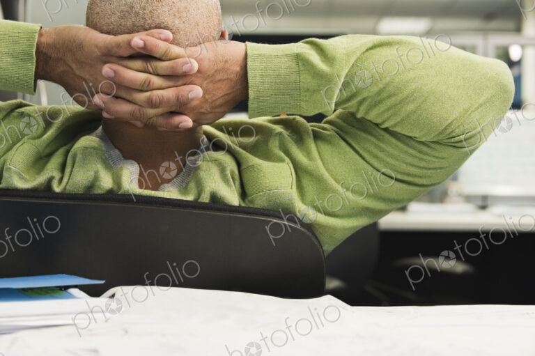 Closeup view from behind of a Black man in a cubicle office with hands behind his head.