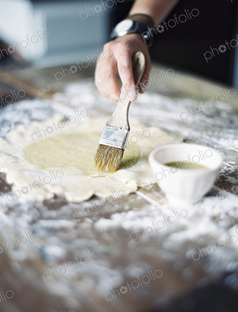 Cook rolling out pastry on a table top and brushing it with egg wash.