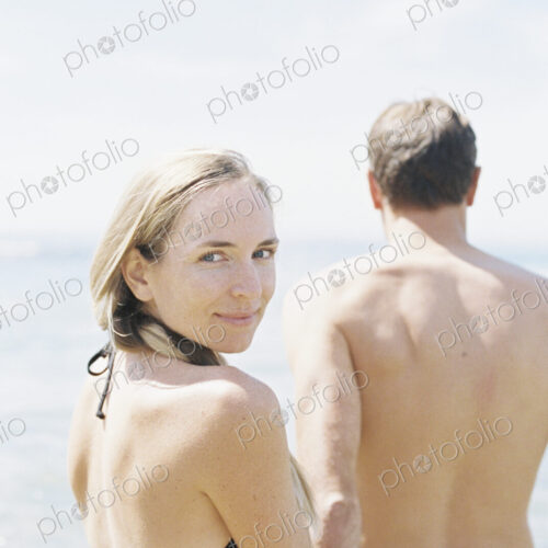 Couple in swimwear by the ocean, woman smiling at camera.