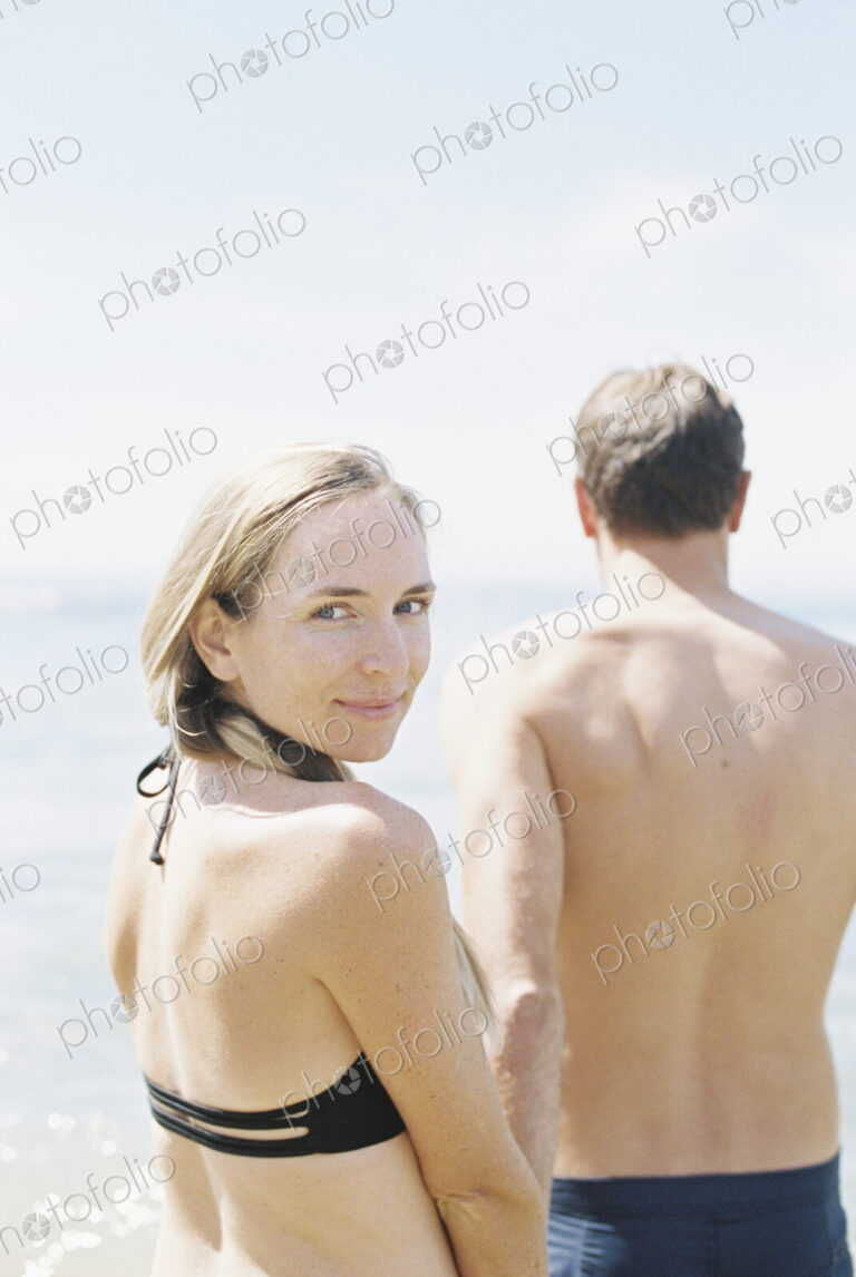 Couple in swimwear by the ocean, woman smiling at camera.