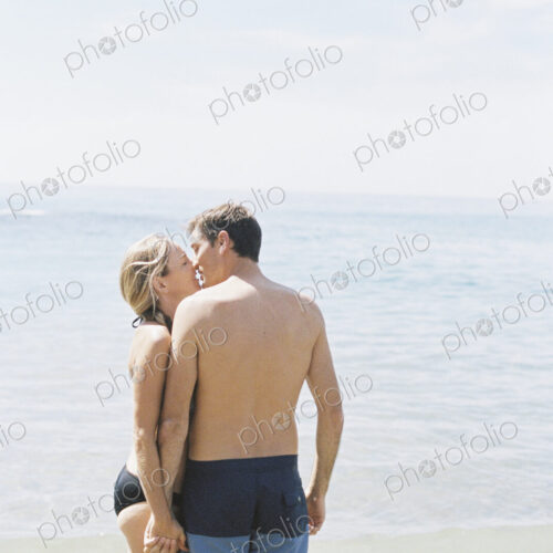 Couple in swimwear standing on a sandy beach by the ocean, kissing.