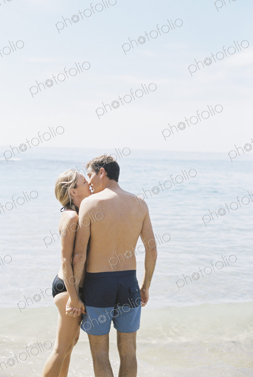 Couple in swimwear standing on a sandy beach by the ocean, kissing.